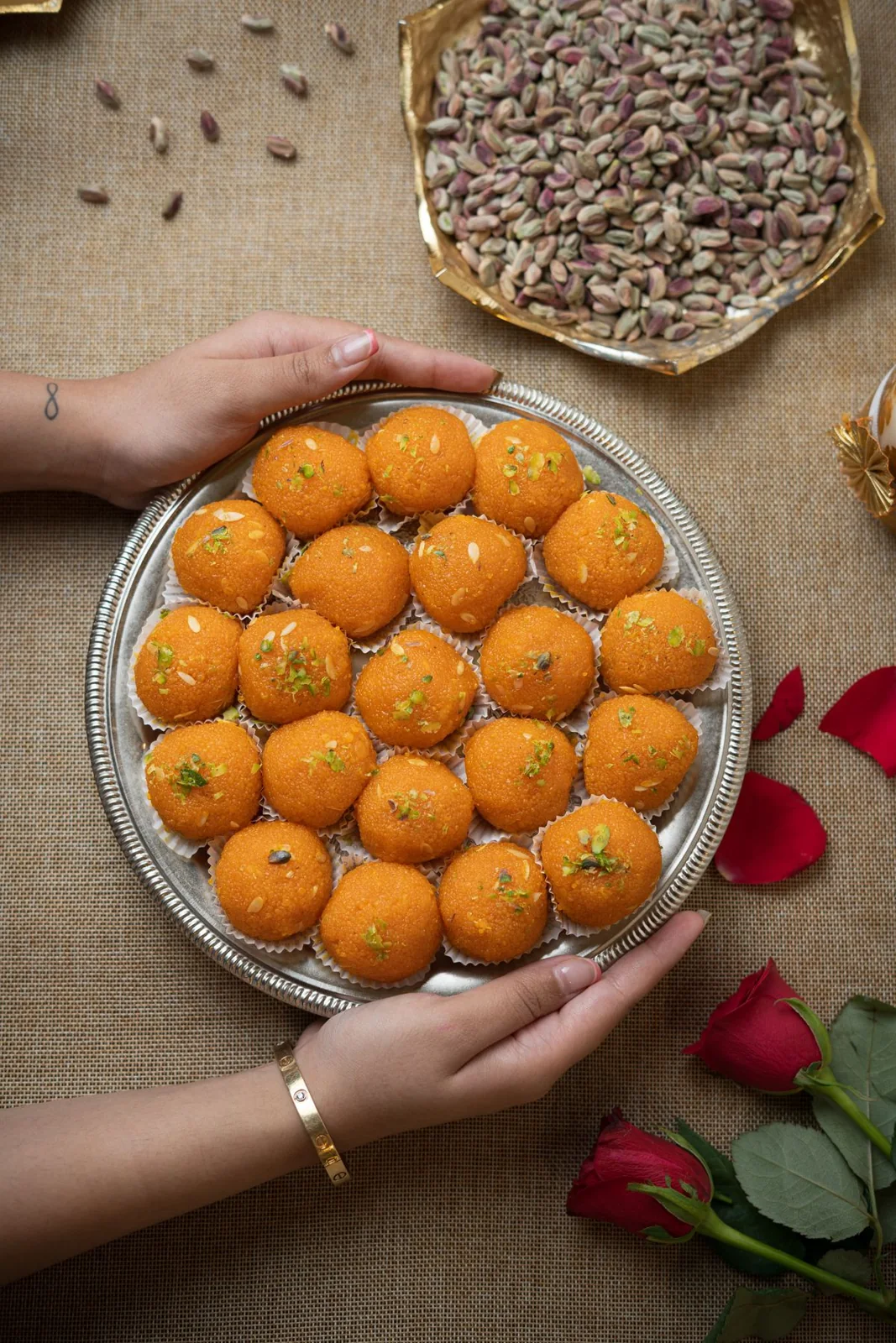Hands holding a tray of laddu sweets beside pistachios and flowers
