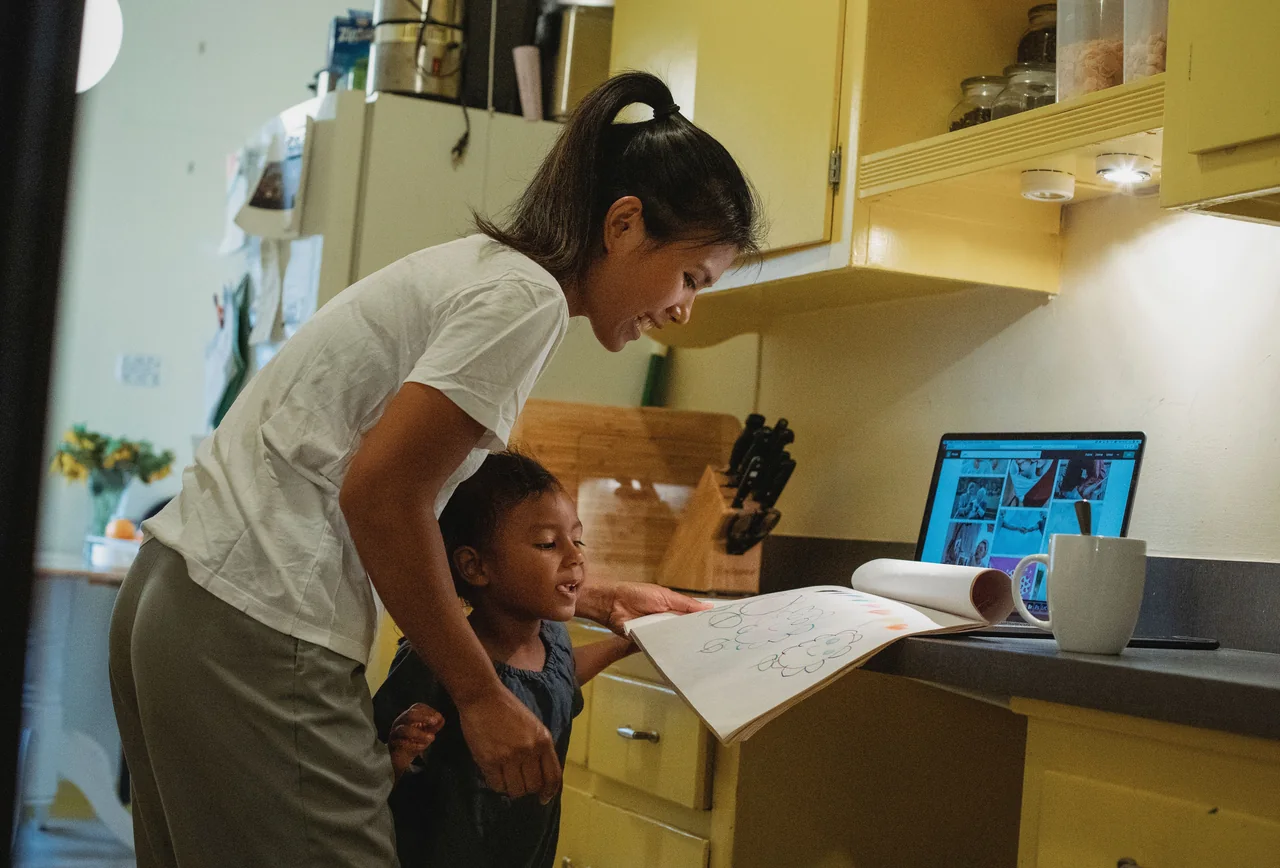 Mother helping her young daughter with a drawing activity beside a laptop