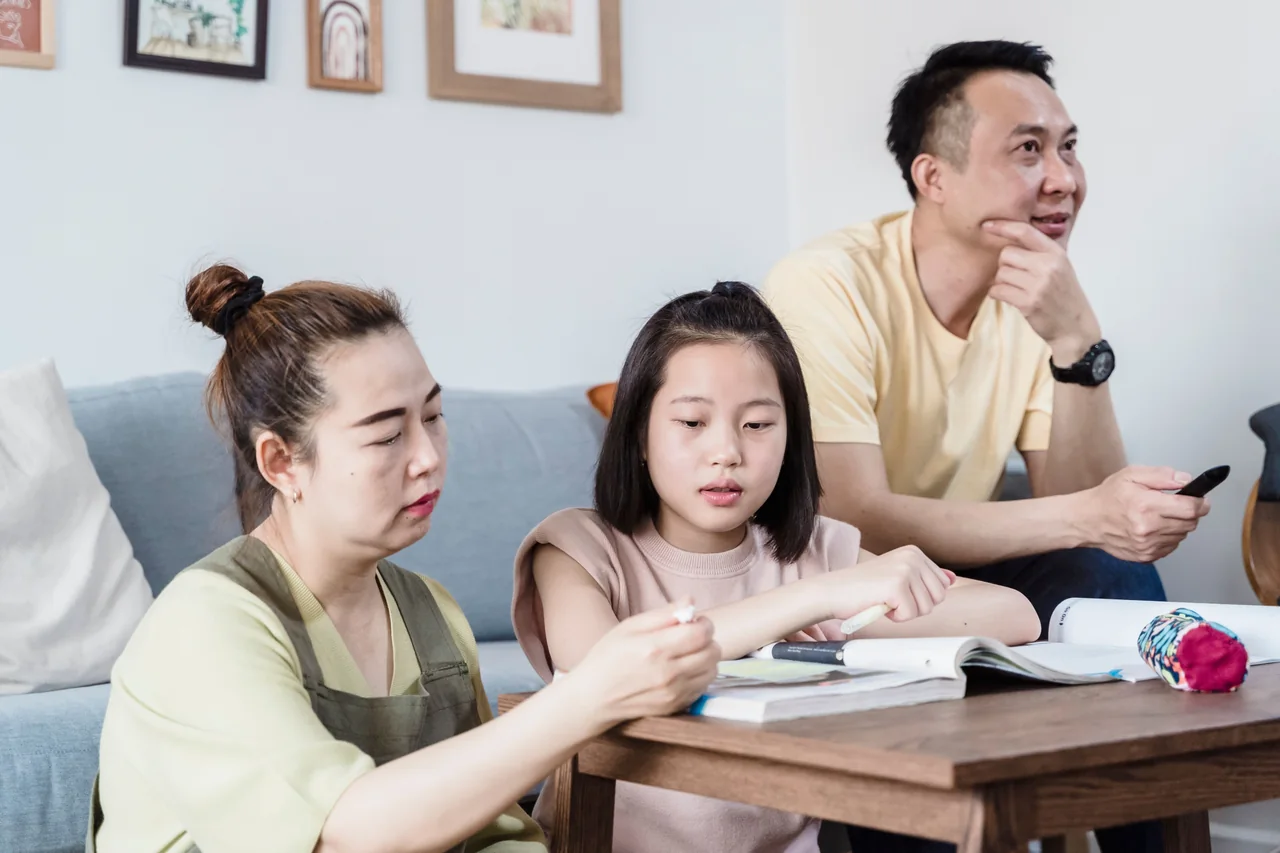 Mother sitting beside her daughter as she writes in a notebook at home
