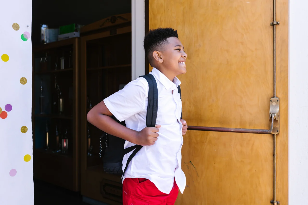 Smiling schoolchild wearing a backpack by a classroom doorway